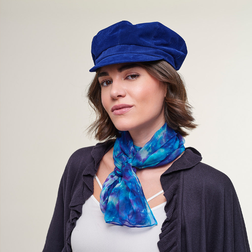A woman wearing a royal blue velvet newsboy cap and a blue and purple patterned silk scarf, styled with a dark cardigan over a white top, against a plain light beige background.