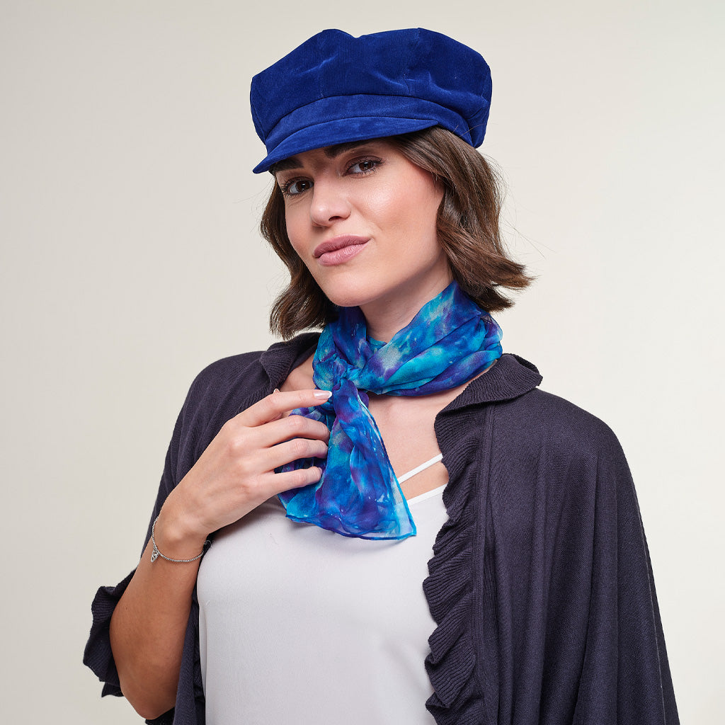A woman wearing a royal blue velvet newsboy cap and a blue and purple patterned silk scarf, styled with a dark cardigan over a white top, against a plain light beige background.