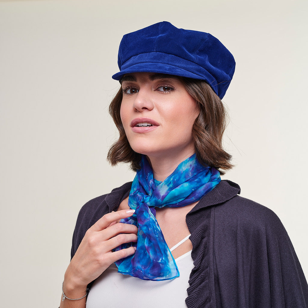 A woman wearing a royal blue velvet newsboy cap and a blue and purple patterned silk scarf, styled with a dark cardigan over a white top, against a plain light beige background.