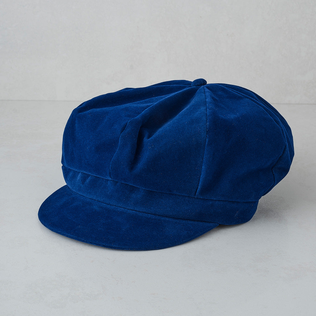 A royal blue velvet newsboy cap with a structured crown and flat brim, photographed on a light gray textured background.
