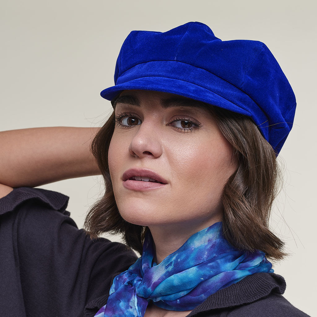 A woman wearing a bright royal blue velvet newsboy cap and a blue and purple galaxy-patterned scarf, photographed against a plain light beige background.