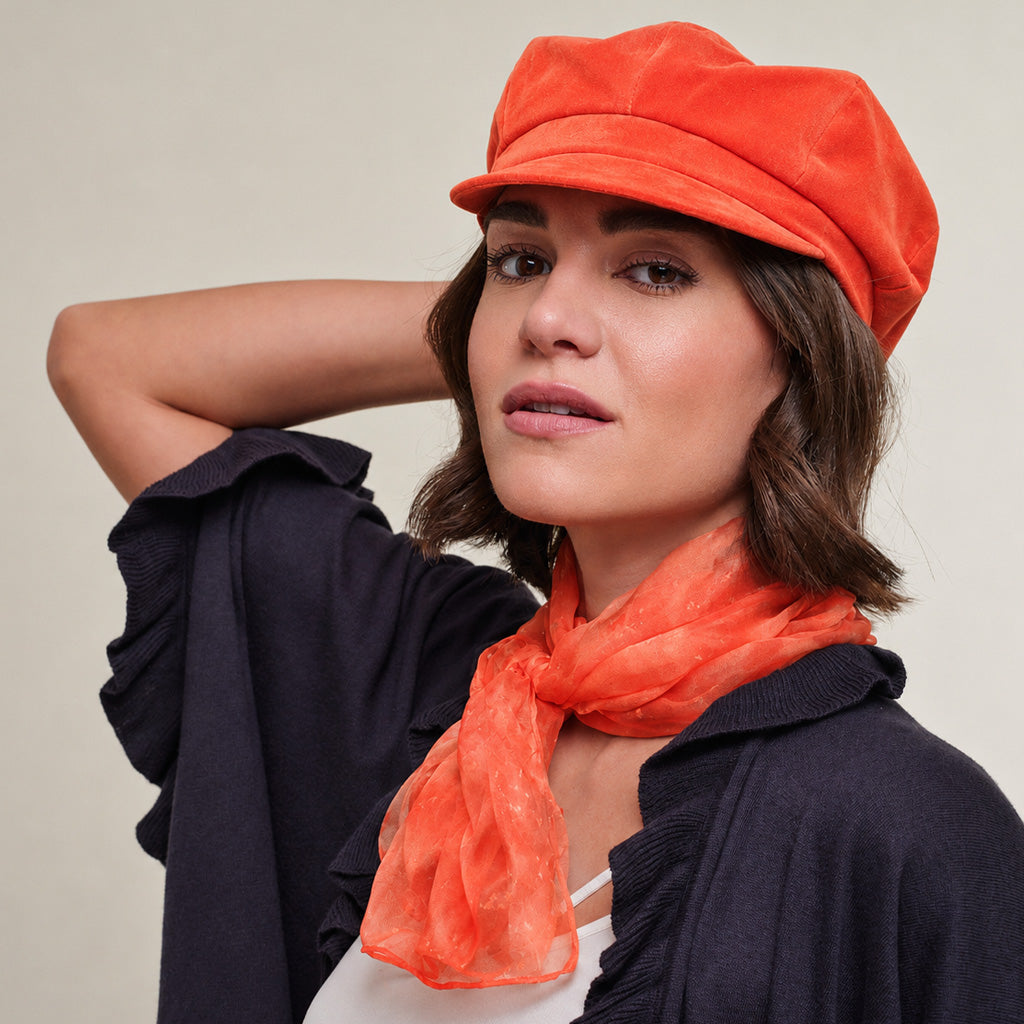 A woman wearing an orange velvet newsboy cap and a matching orange scarf, paired with a dark navy cardigan, set against a plain beige background.