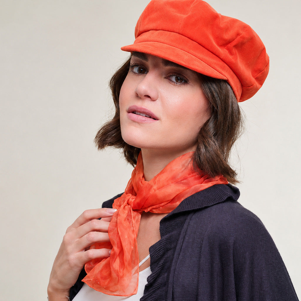 A woman wearing an orange velvet newsboy cap and a matching orange scarf, paired with a dark navy cardigan, set against a plain beige background.