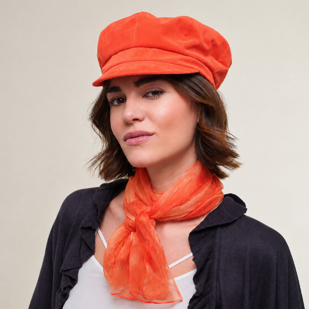A woman wearing an orange velvet newsboy cap and a matching orange scarf, paired with a dark navy cardigan, set against a plain beige background.