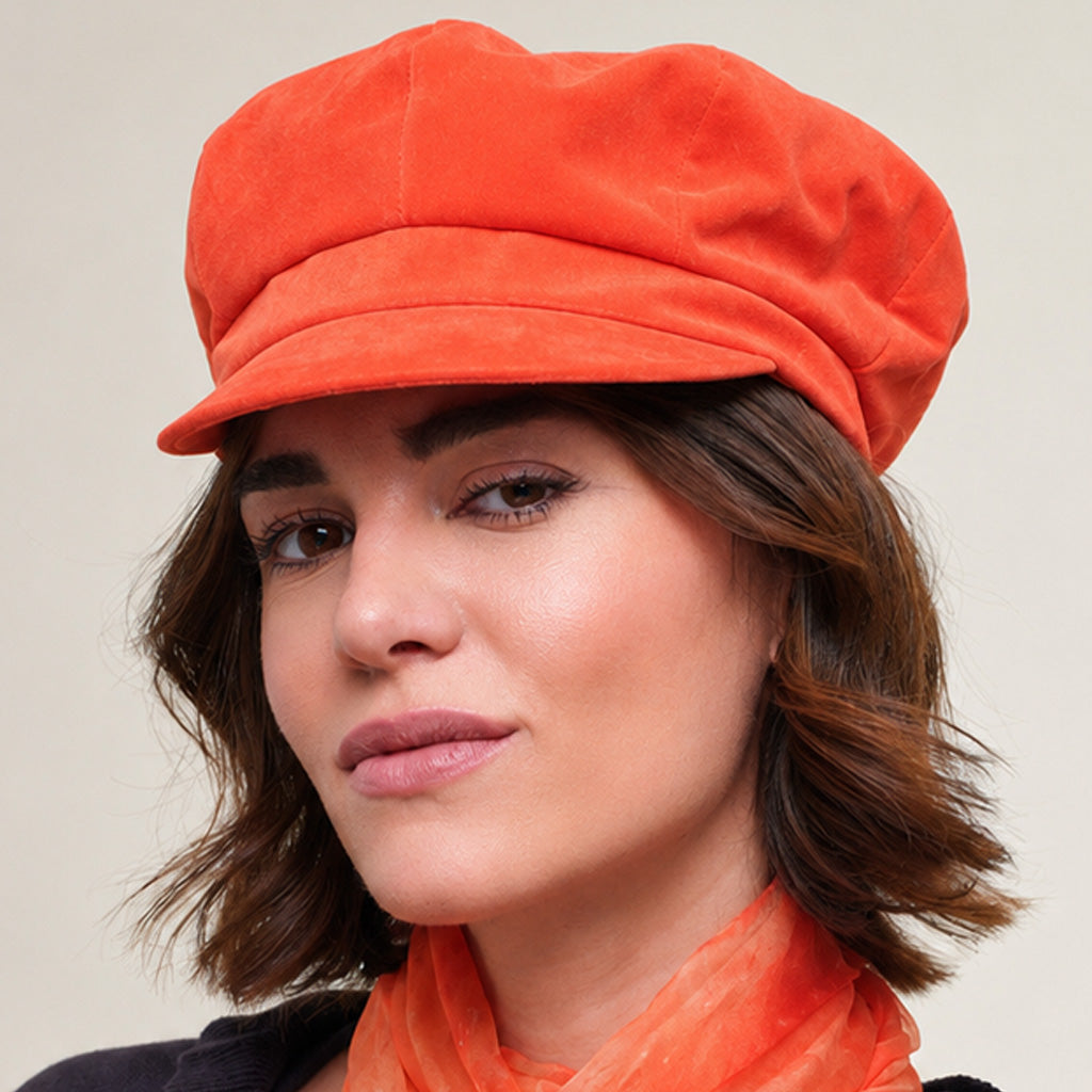 A woman wearing an orange velvet newsboy cap and a matching orange scarf, paired with a dark navy cardigan, set against a plain beige background.