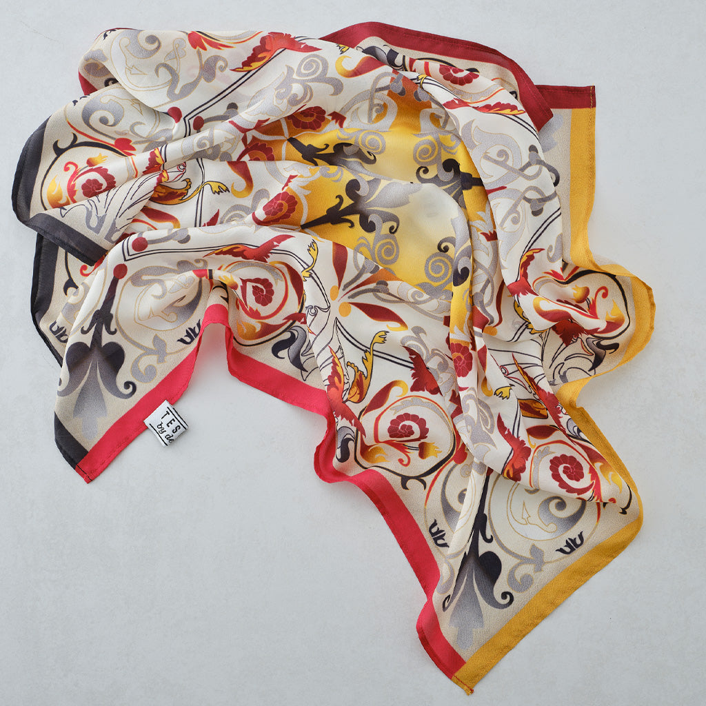 A square silk scarf featuring a symmetrical, ornate folk-art style design with red roses, yellow sunburst, and black, gray, and gold details on a cream background. The brand name 'TECCA' is printed in the center.