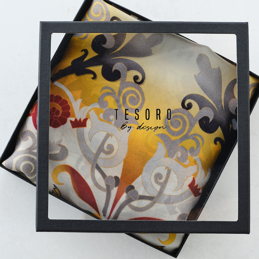 A square black-framed glass or acrylic jewelry box lid with a clear center panel, shown open to reveal a decorative silk scarf inside. The scarf has an ornate baroque-style pattern in gold, gray, black, red, and cream, with the text 'TESORO by design' printed on it.