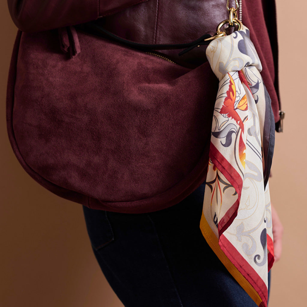 A close-up of a person wearing a burgundy suede shoulder bag with a patterned silk scarf hanging from the zipper pull. The person is also wearing dark blue jeans, set against a solid warm brown background.