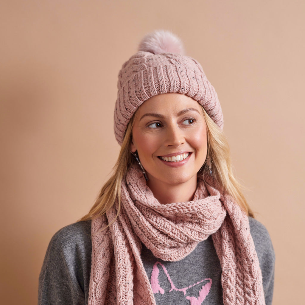 A smiling woman wearing a dusty pink cable-knit beanie with a matching pom-pom and a chunky knit scarf, paired with a gray sweater against a solid beige background.