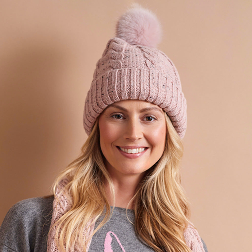A smiling blonde woman wearing a dusty pink cable-knit beanie with a matching pom-pom and a pink knit scarf, set against a solid warm beige background.