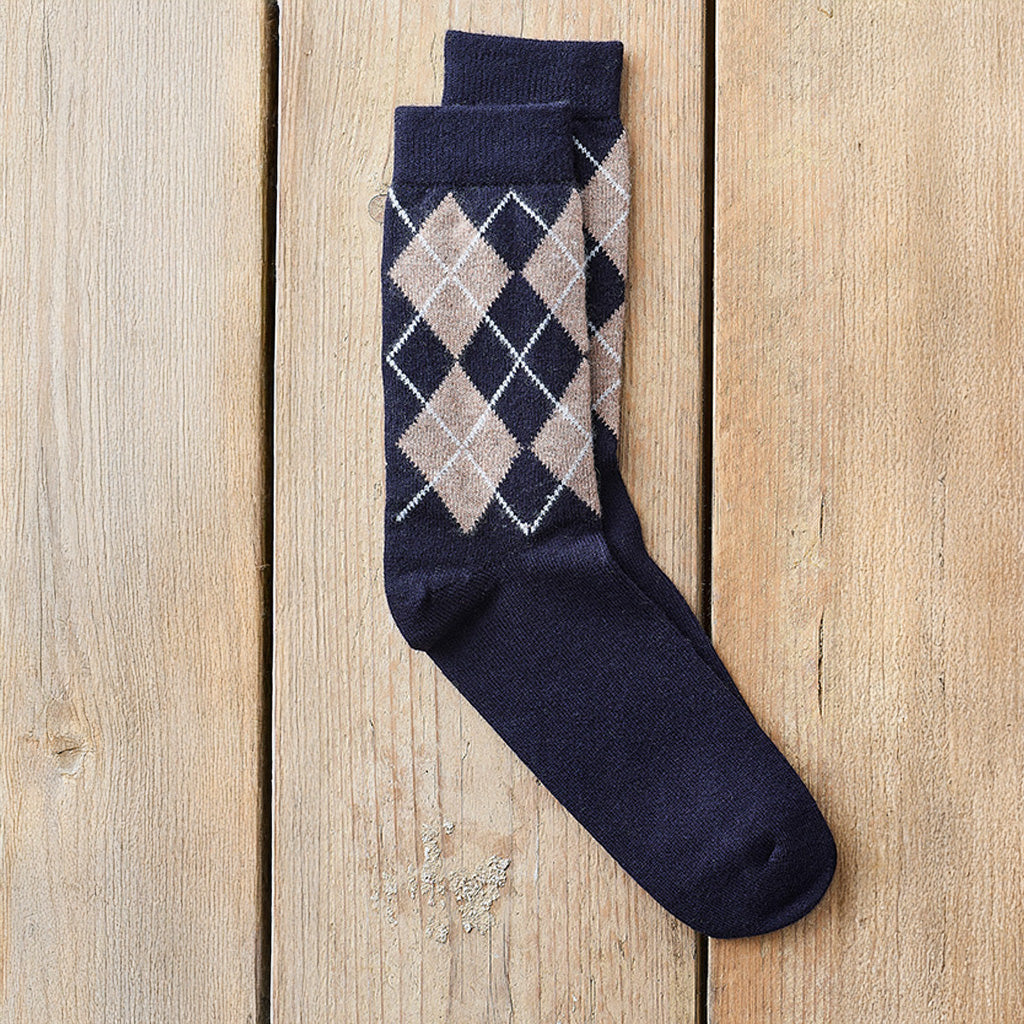 A single navy blue crew sock with an argyle diamond pattern in beige, white, and navy, laid flat on a light wood plank background.