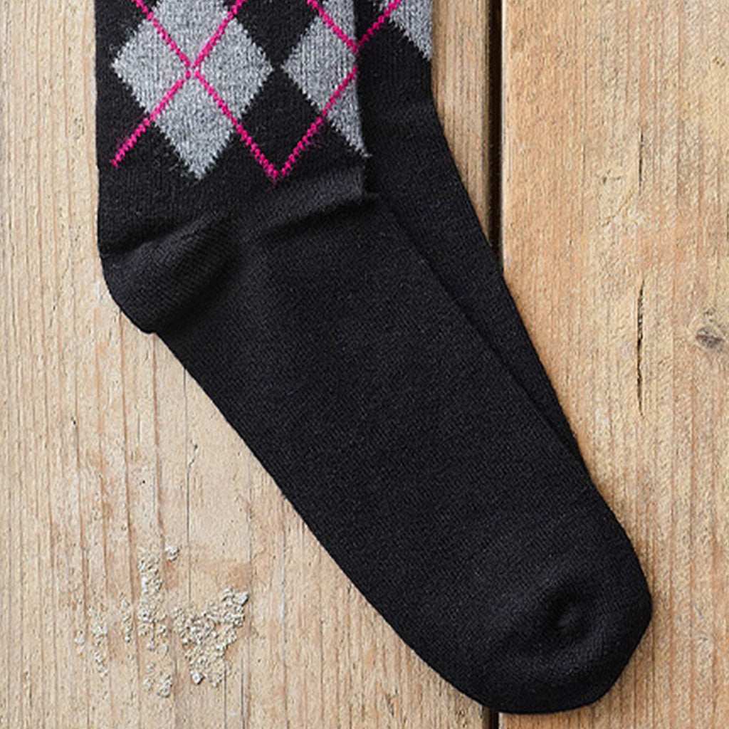 A single black knit sock with a gray and black argyle pattern outlined in pink near the cuff, laid diagonally on a light wood plank surface.