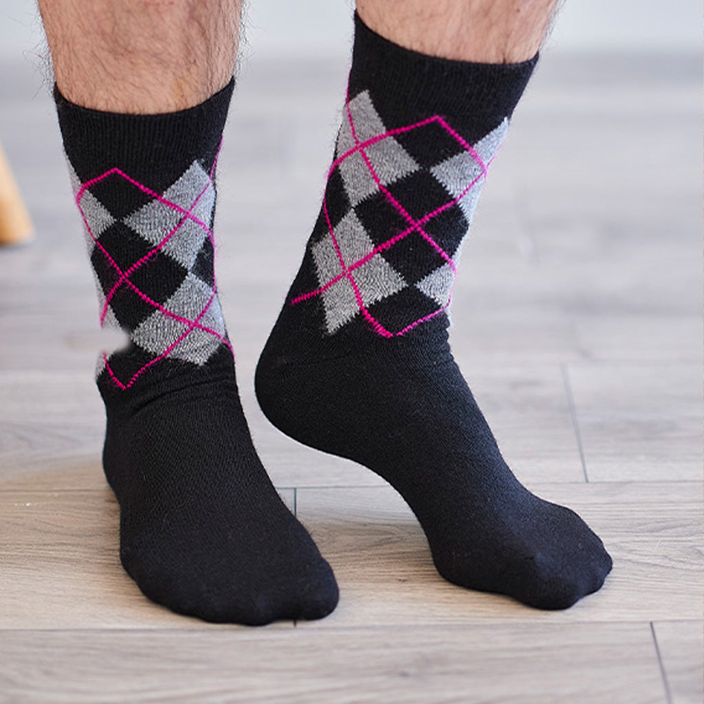 A pair of black crew socks with a gray and pink argyle pattern on the upper calf, worn on a person's feet on a light wood floor.
