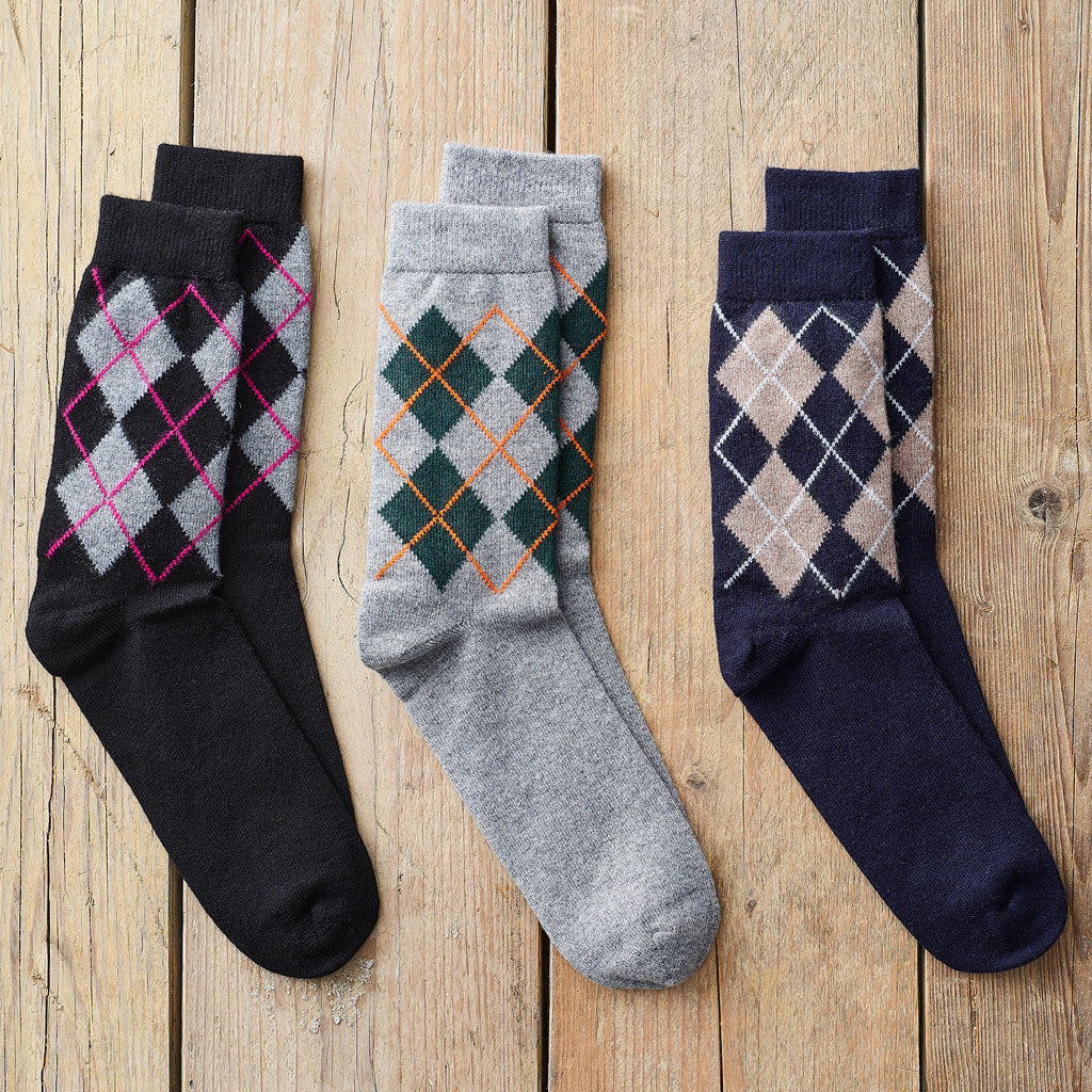Three pairs of men's crew socks with argyle patterns, laid flat on a light brown wooden plank background. The left pair is black with a pink and gray argyle pattern.