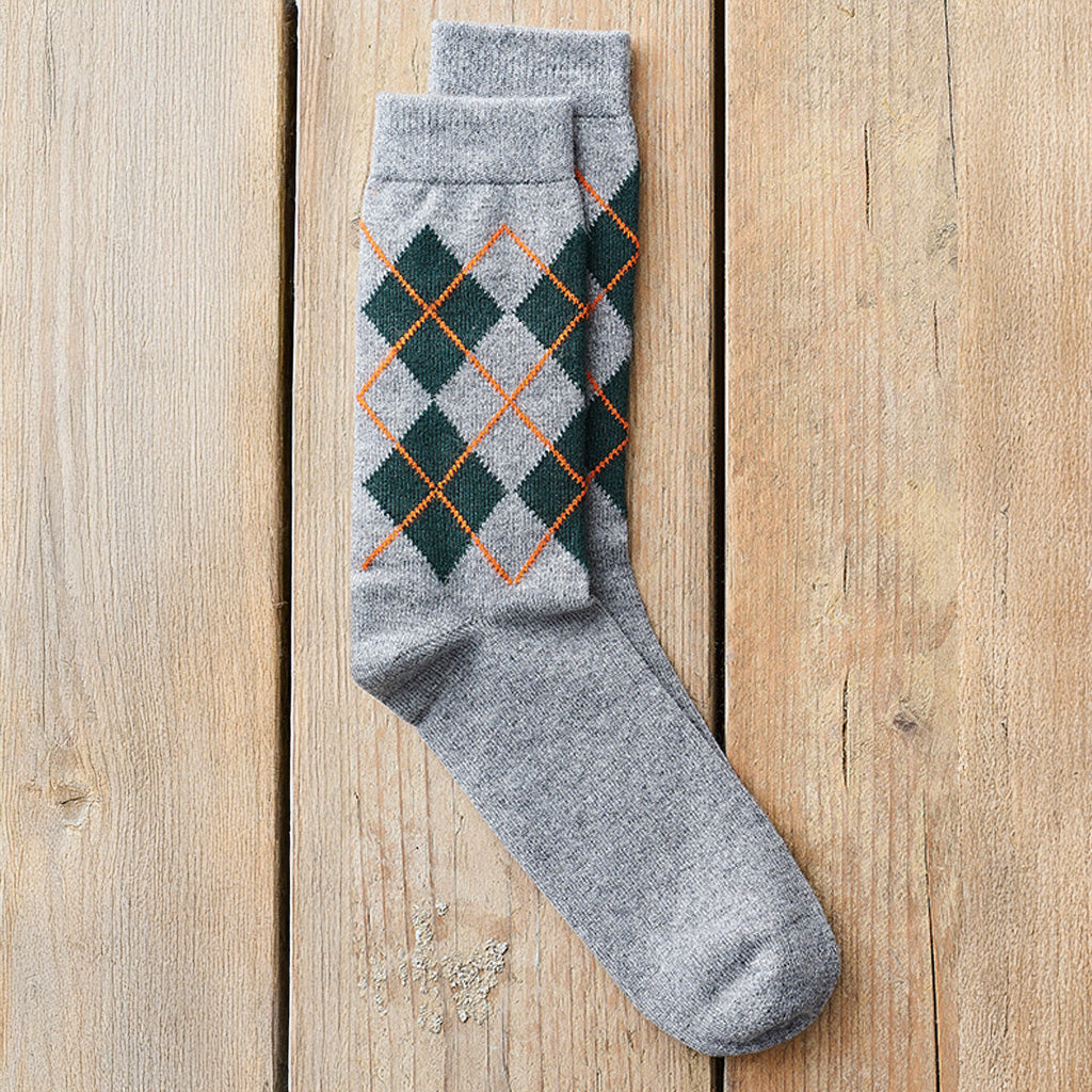 A single gray knit sock with an argyle pattern in dark green and orange, laid flat on a light wood plank background.