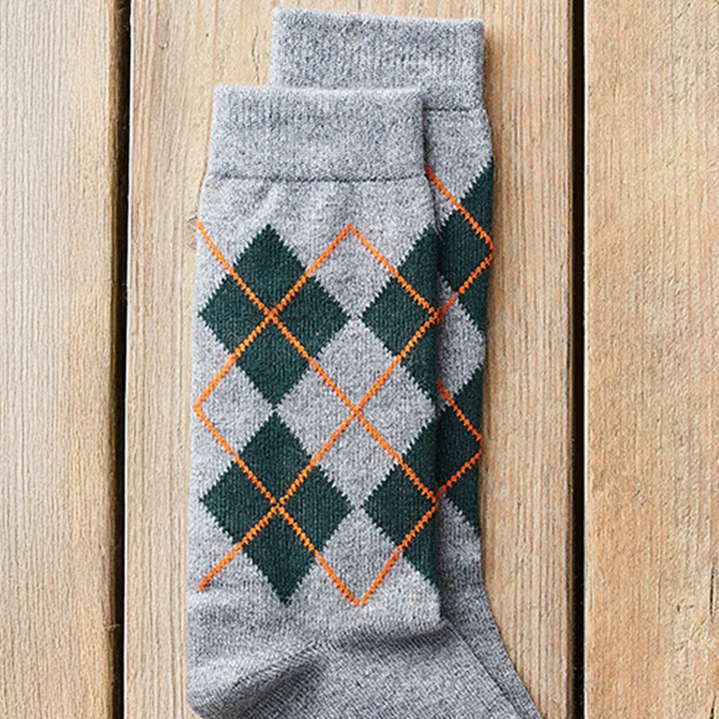 A pair of gray knit socks with an argyle pattern in dark green and orange, laid flat on a light wood plank background.