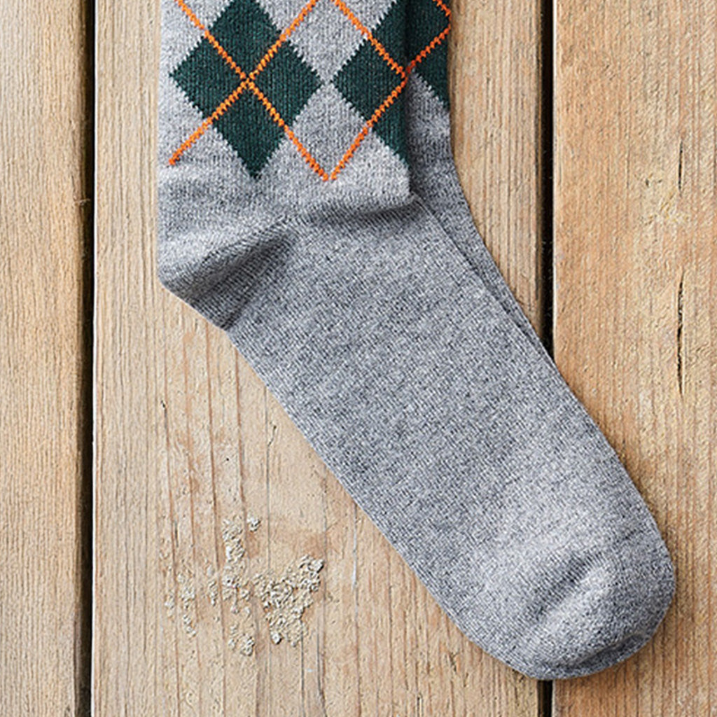 A single gray knit sock with an argyle pattern in dark green, light gray, and orange, laid diagonally on a light brown wooden plank surface.