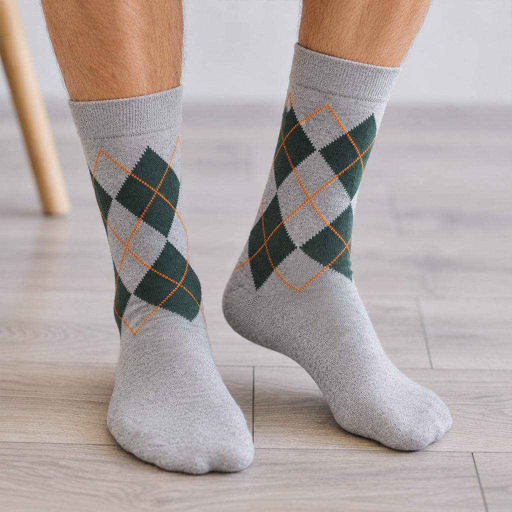 A pair of gray crew socks with a green and orange argyle pattern, worn on a person's feet on a light wood floor.