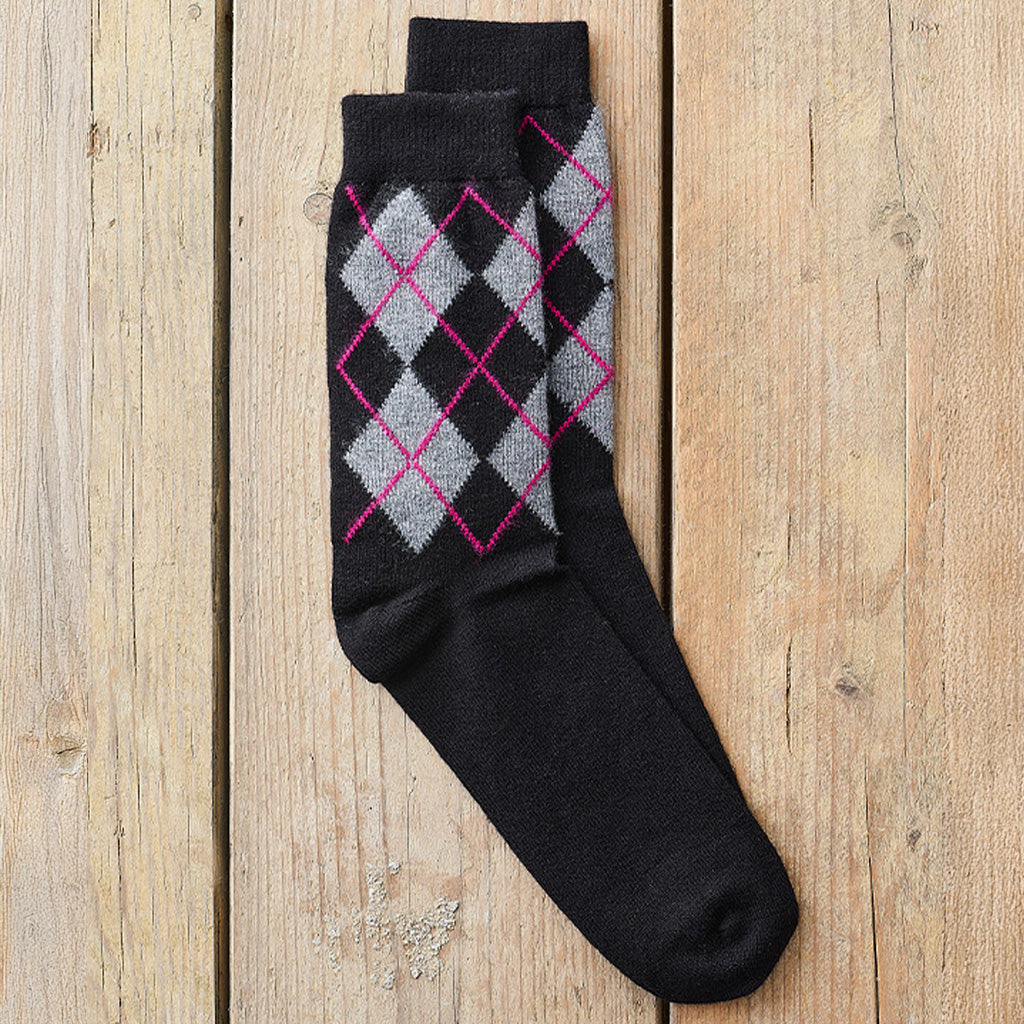 A pair of black crew socks with a gray and pink argyle pattern on the upper leg, laid flat on a light brown wooden plank background.