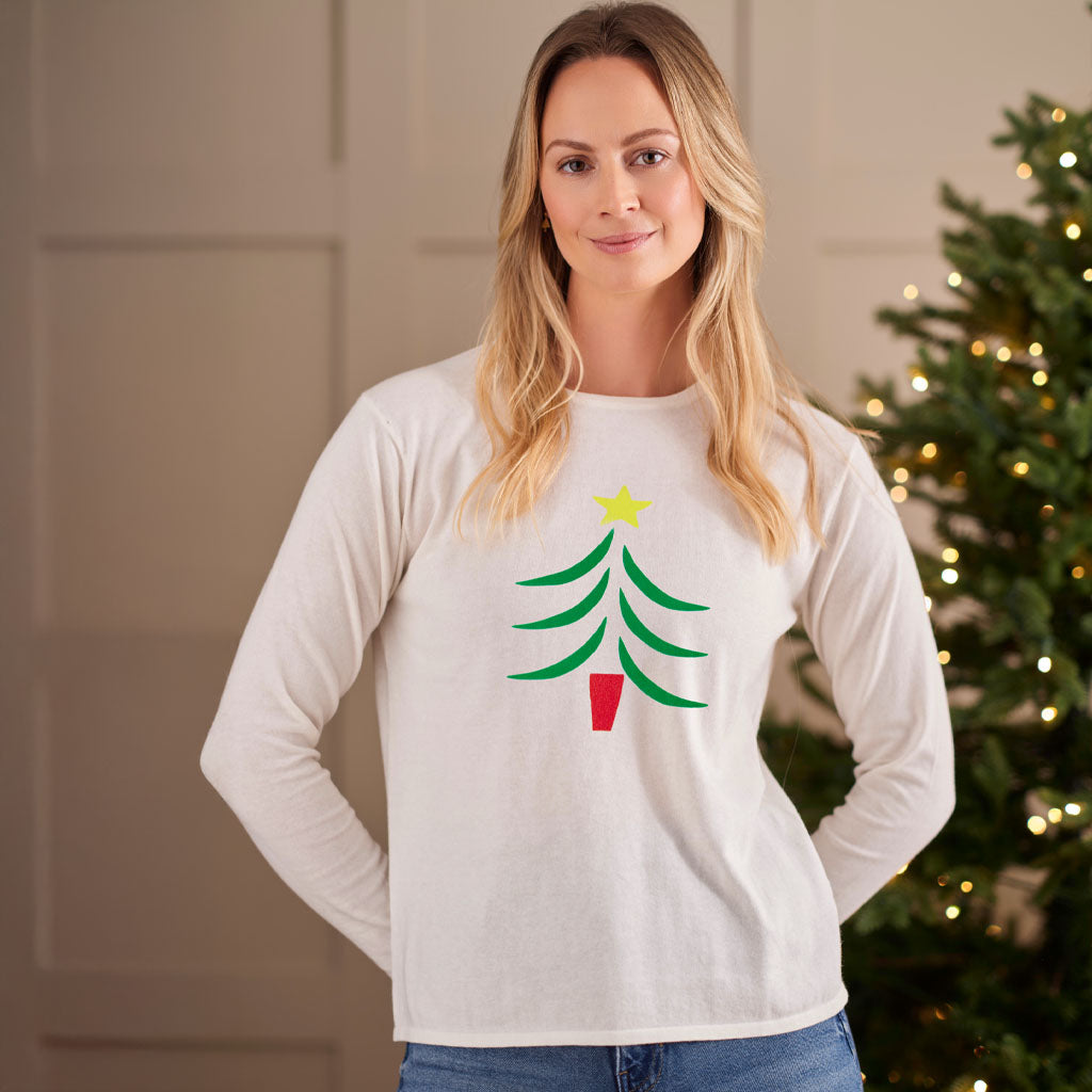 A woman wearing a white long-sleeve crewneck shirt with a green Christmas tree graphic and a yellow star on the front, posed in front of a softly lit holiday tree.