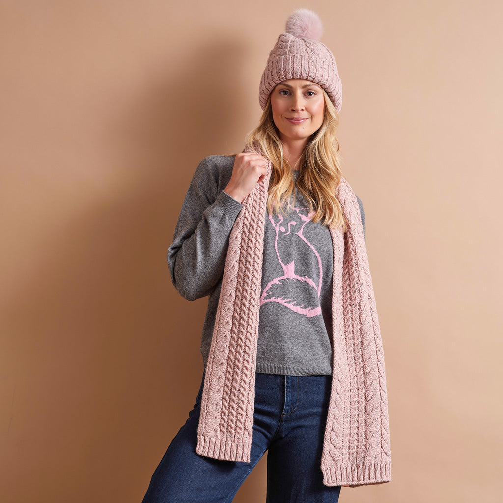 A woman modeling a light pink cable-knit beanie with a pom-pom, a matching pink cable-knit scarf, and a gray graphic sweater, posed against a solid warm beige background.