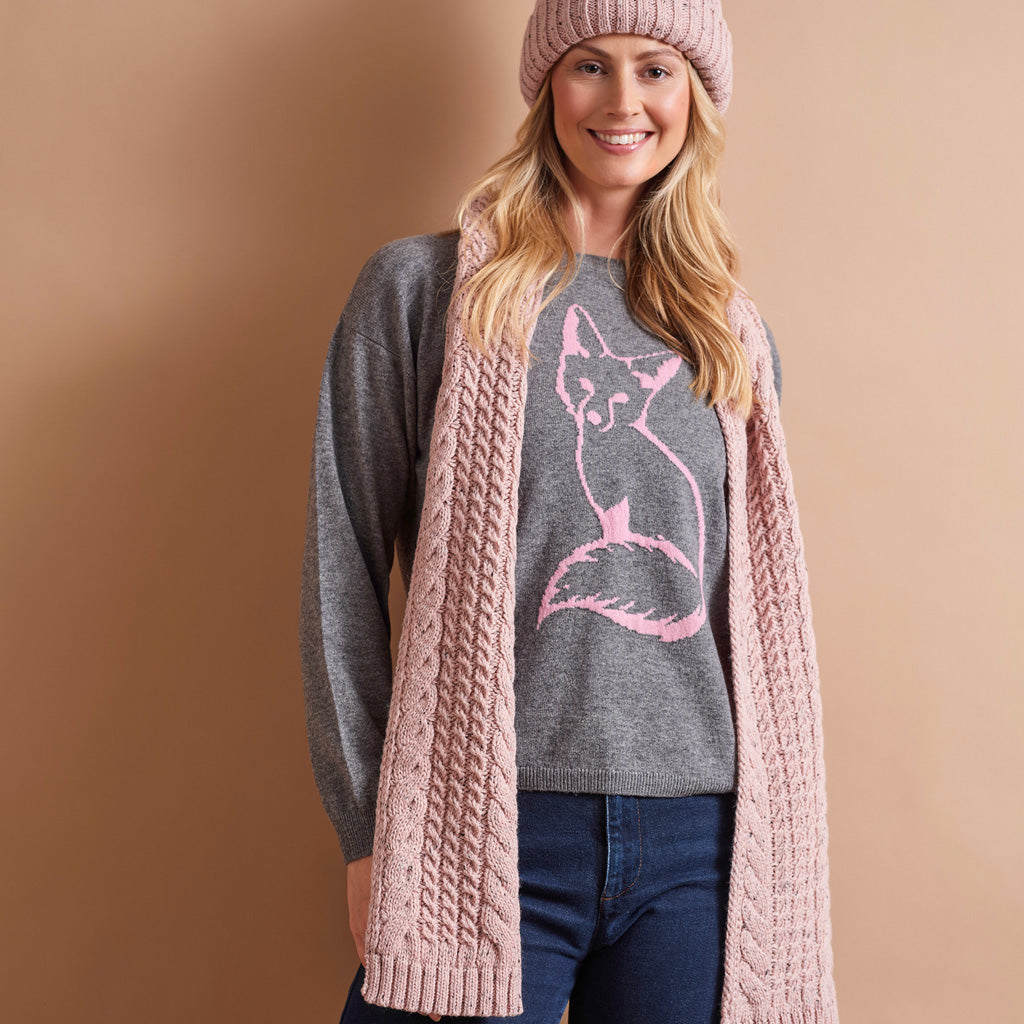 A woman wearing a chunky pink cable-knit cardigan over a heather gray crewneck sweater with a pink fox graphic, paired with a pink knit beanie and blue jeans, set against a solid warm tan background.