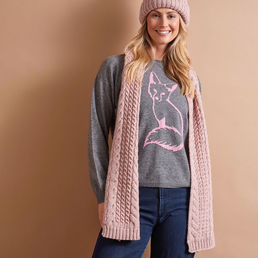 A woman wearing a pink cable-knit cardigan over a gray crewneck sweater with a pink cat graphic, paired with blue jeans and a pink knit beanie, set against a solid tan studio background.