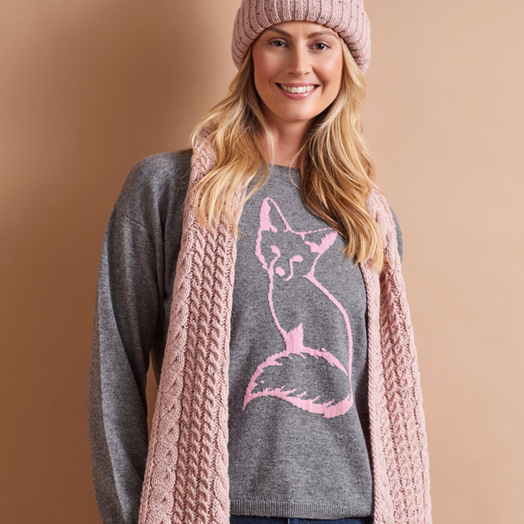A smiling woman wearing a pink knit beanie, a gray sweater with a pink fox graphic, and a chunky pink cable-knit cardigan, posed against a solid warm beige background.