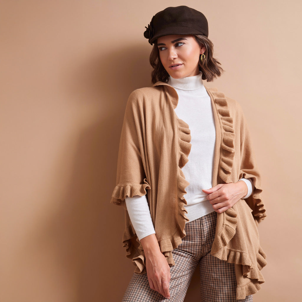 A woman models a camel-colored ruffled cardigan over a white turtleneck, paired with brown and white checkered trousers and a dark brown newsboy cap, set against a solid beige background.