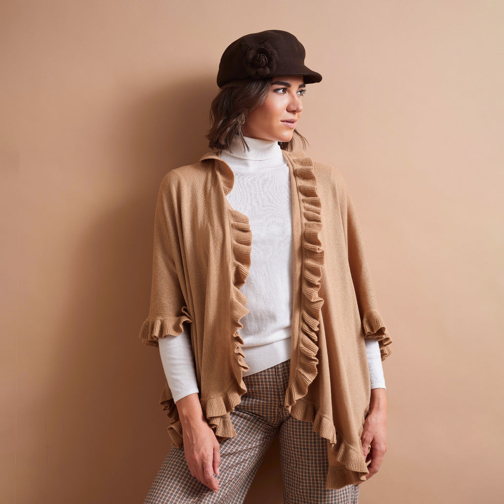 A woman models a camel brown ruffled cape cardigan over a white turtleneck sweater, paired with brown and beige check trousers and a dark brown felt hat, set against a solid warm beige background.