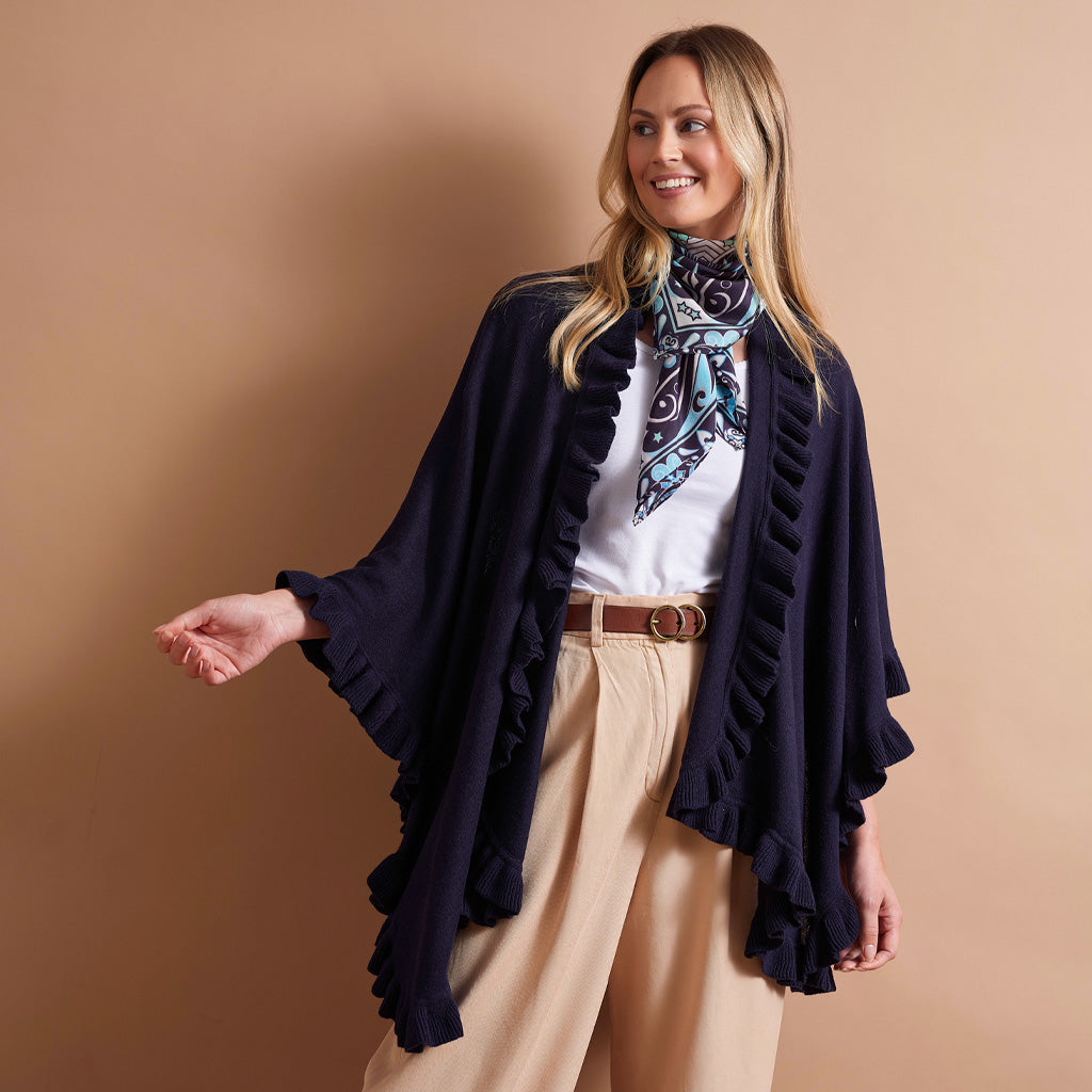 A woman modeling a navy blue ruffled open-front cardigan over a white top and beige pants, accessorized with a patterned scarf, against a solid tan studio background.