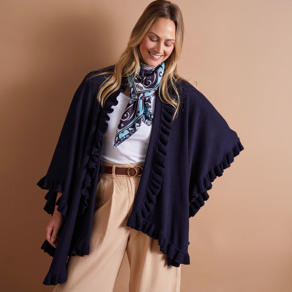 A woman modeling a navy blue open-front poncho with ruffled trim, worn over a white top and beige trousers, with a patterned scarf, against a solid tan studio background.