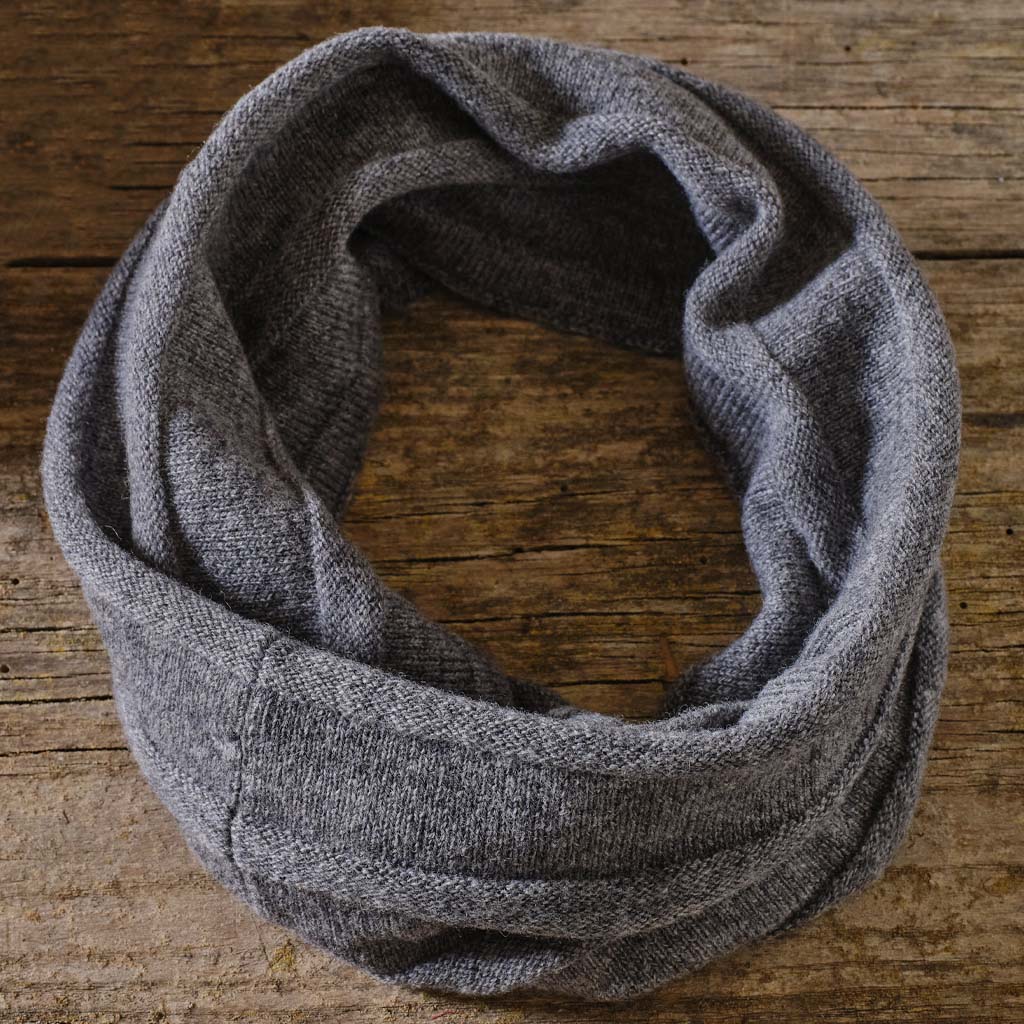A gray knit infinity scarf with a textured herringbone pattern, arranged in a loose loop on a rustic wooden surface.