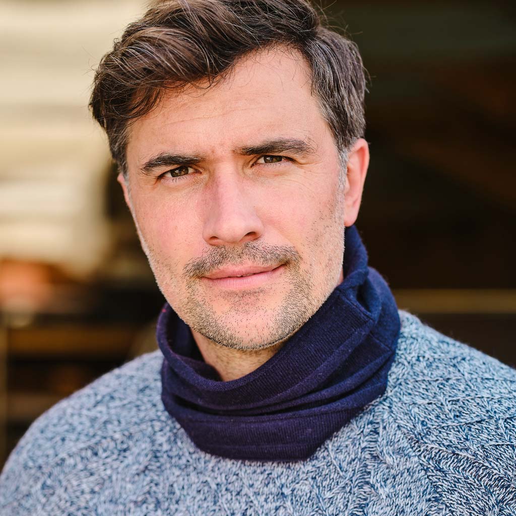 A close-up portrait of a man with short brown hair and light stubble, wearing a dark navy knit turtleneck sweater layered under a heathered light blue knit sweater, against a softly blurred warm-toned background.