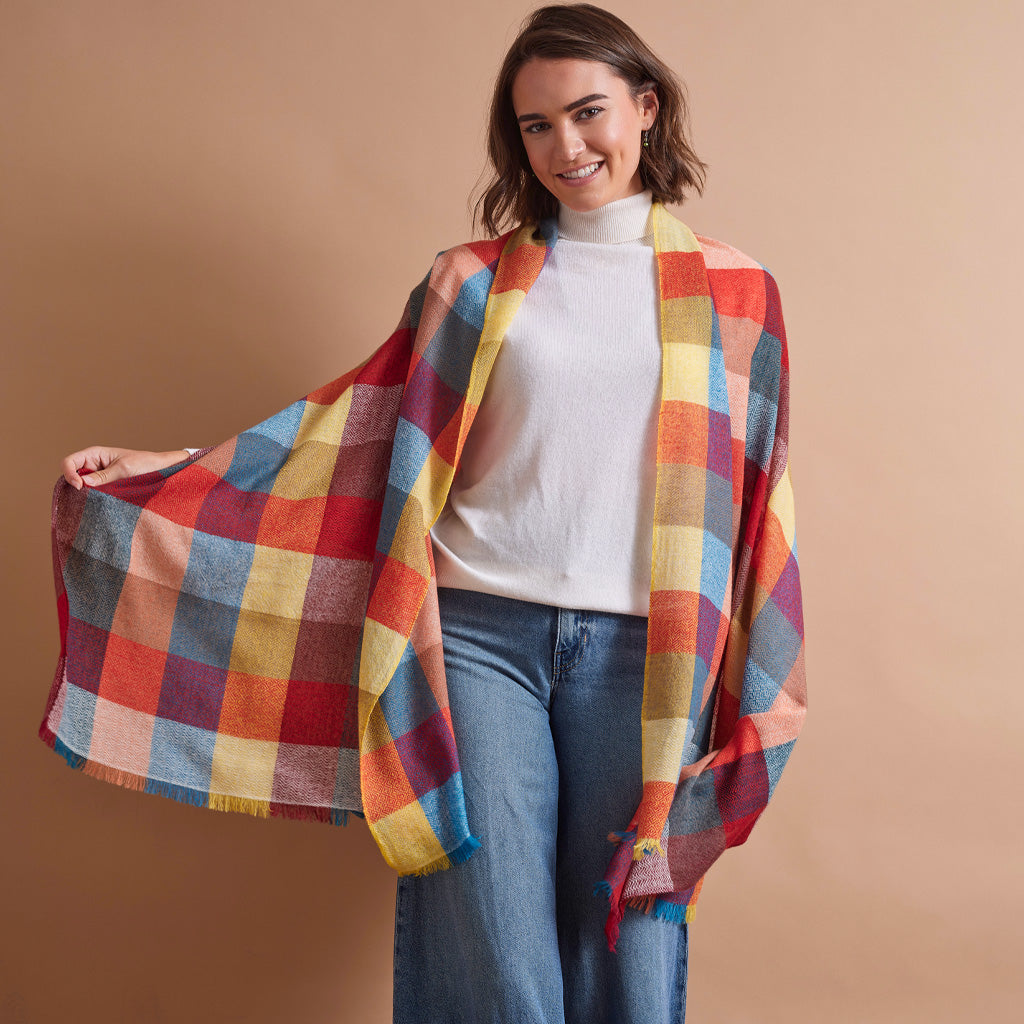A woman modeling a vibrant multicolor plaid wrap shawl draped over her shoulders, paired with a white turtleneck and blue jeans, set against a solid warm beige studio background.