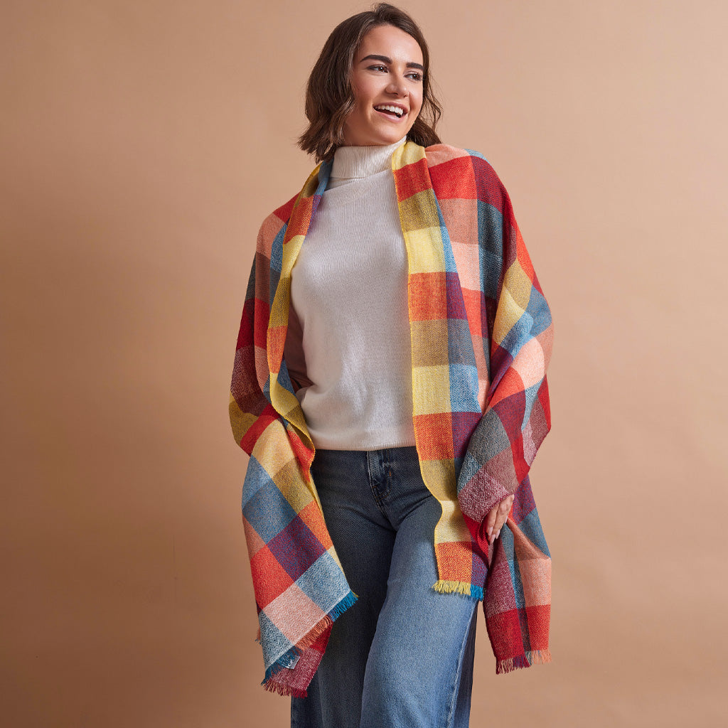 A woman smiling and wearing a vibrant, multicolor plaid wrap shawl over a light gray turtleneck sweater and blue jeans, set against a solid warm beige studio background.