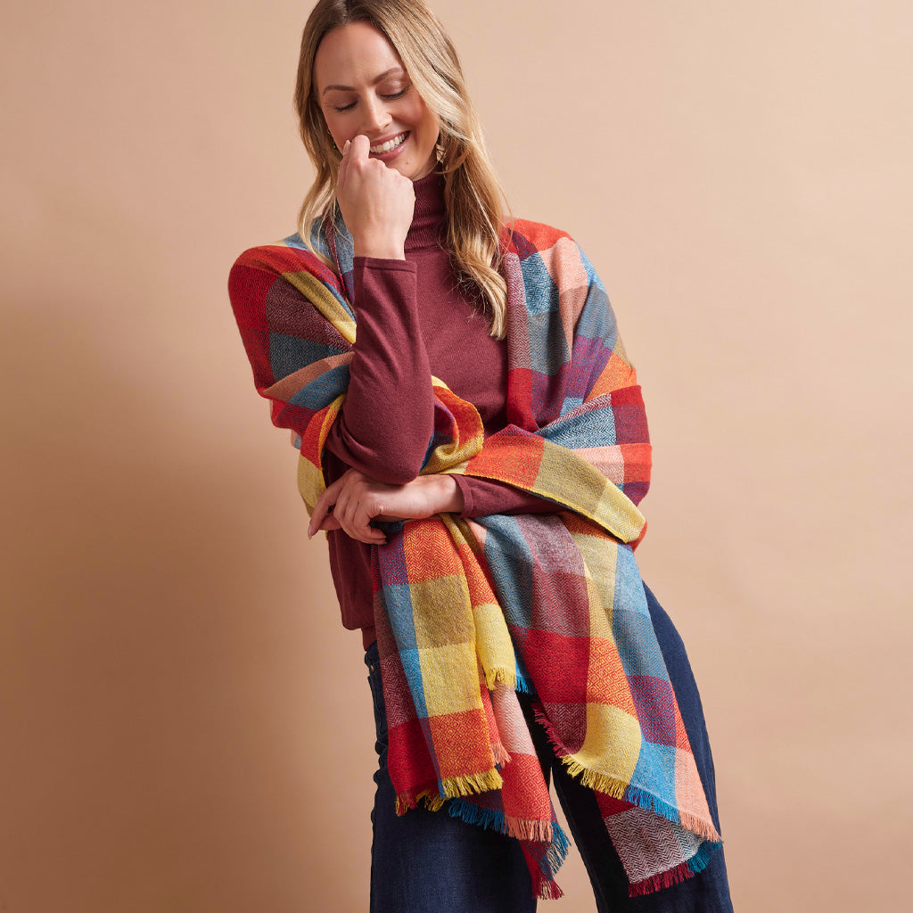 A woman wearing a multicolor plaid shawl over a burgundy turtleneck and dark jeans, posing against a solid beige studio background.