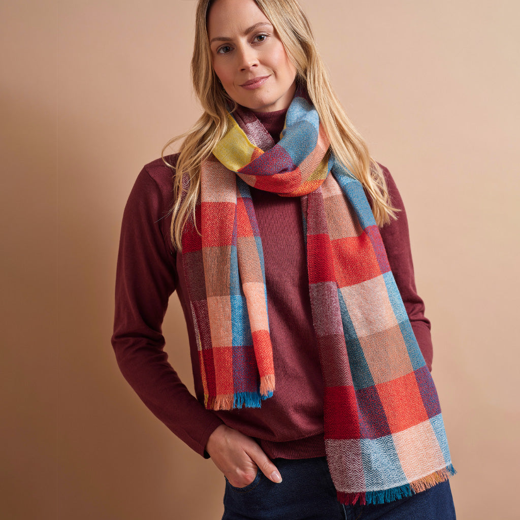 A woman wearing a multicolor plaid scarf with red, blue, yellow, and beige tones, styled over a burgundy sweater and dark jeans against a solid beige background.