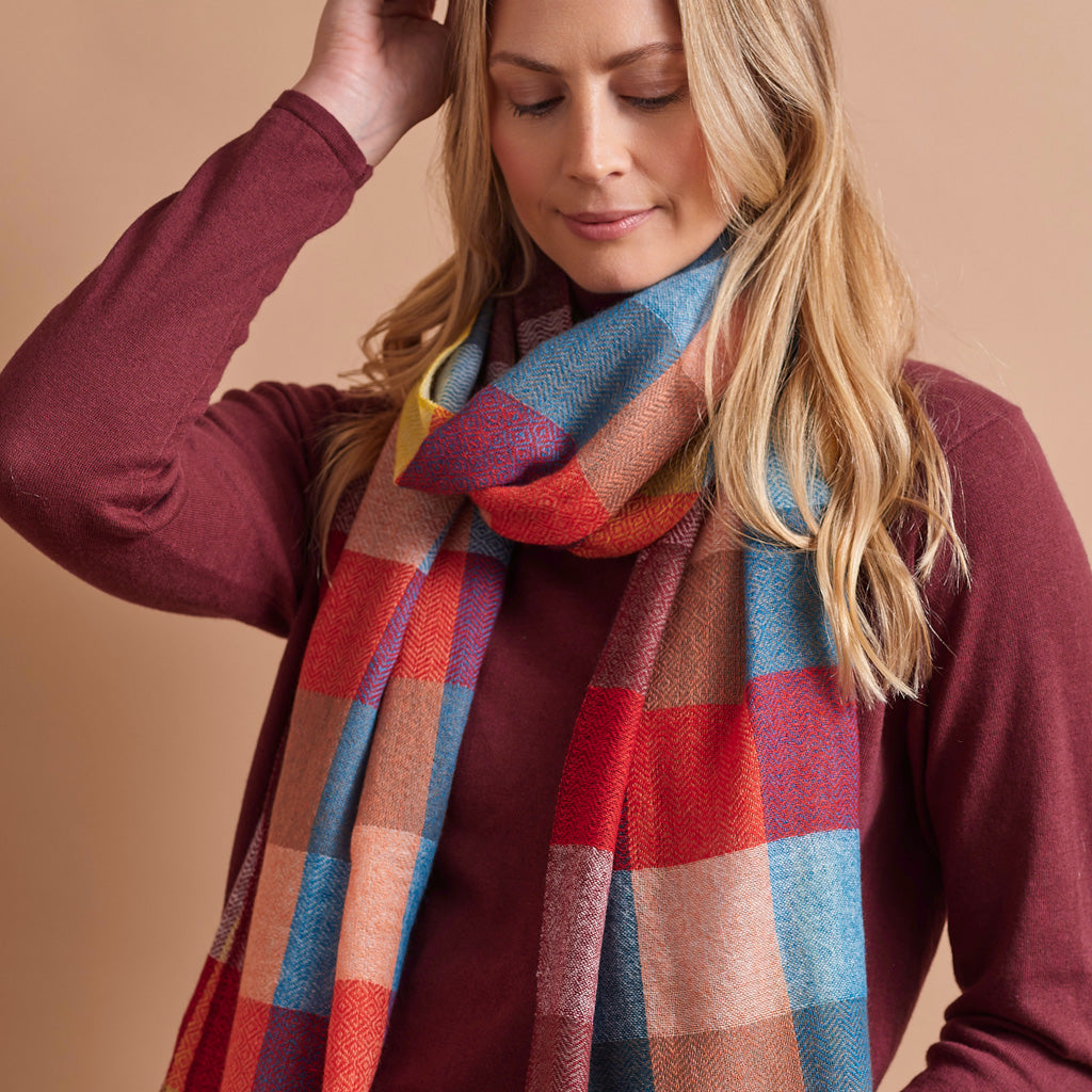 close up A woman modeling a vibrant multicolor plaid wrap shawl styled around her neck wearing turtleneck and blue jeans, set against a solid warm beige studio background.
