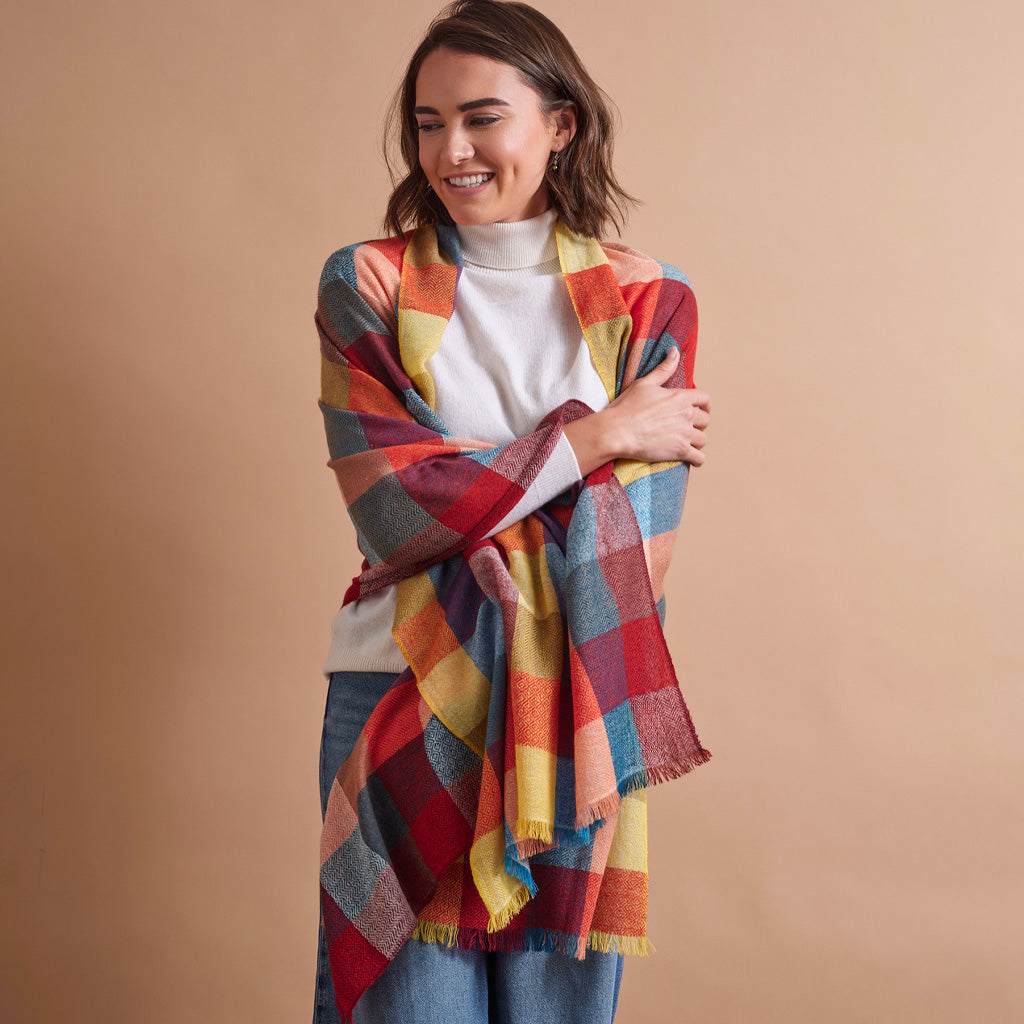 A woman wearing a white turtleneck and blue jeans is wrapped in a large, multicolor plaid scarf with red, yellow, blue, and orange tones, set against a solid beige background.
