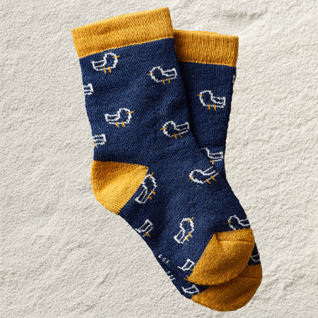 A pair of navy blue crew socks with mustard yellow cuffs, heels, and toes, featuring a white and yellow bird pattern, laid flat on a textured light beige surface.