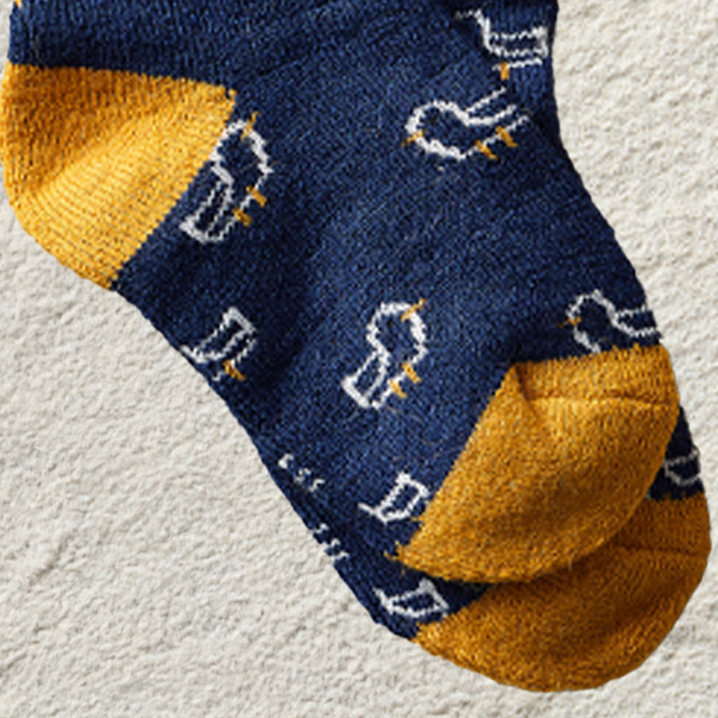 A close-up of a single knit sock in navy blue with mustard yellow toe and cuff sections, plus small white and yellow duck motifs knitted across the foot, shown on a light textured surface.
