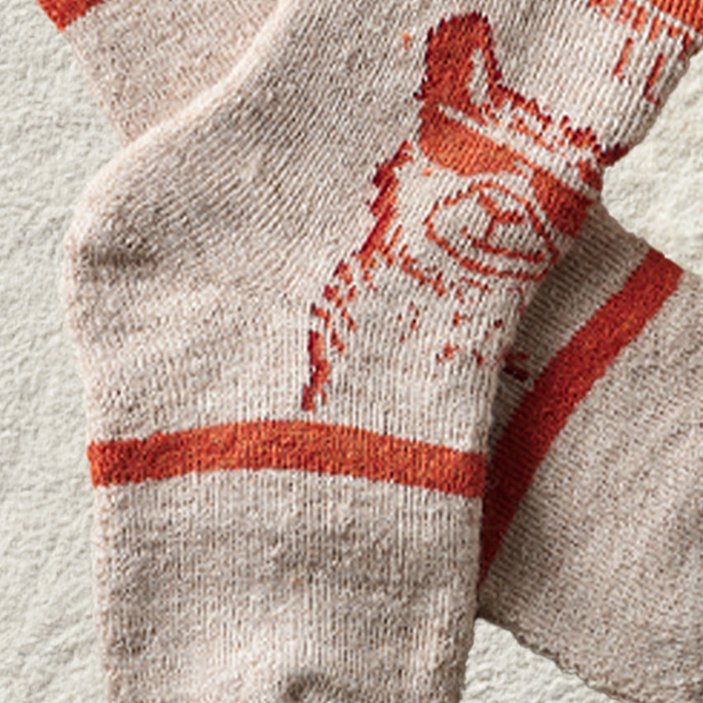 Close-up of a pair of knit socks in a light heathered beige or off-white color with a bold orange-red graphic and stripe details.