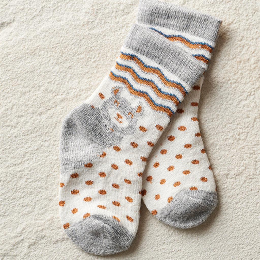 A pair of baby socks with a white base, orange polka dots, a gray heel and toe, and blue and orange wavy stripes near the cuff, displayed on a textured beige fabric background.