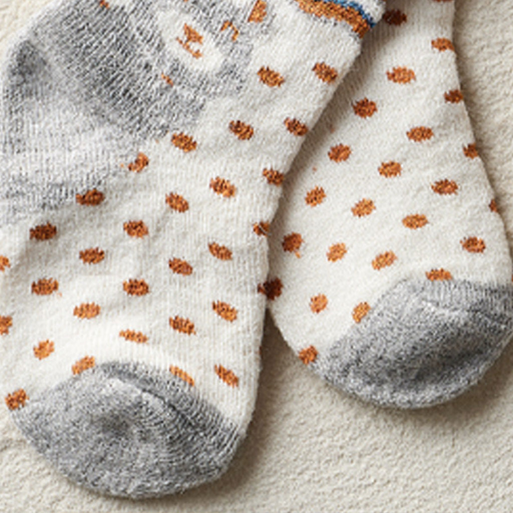 A pair of white knit socks with orange polka dots and gray toes and heels, laid flat on a textured beige surface.