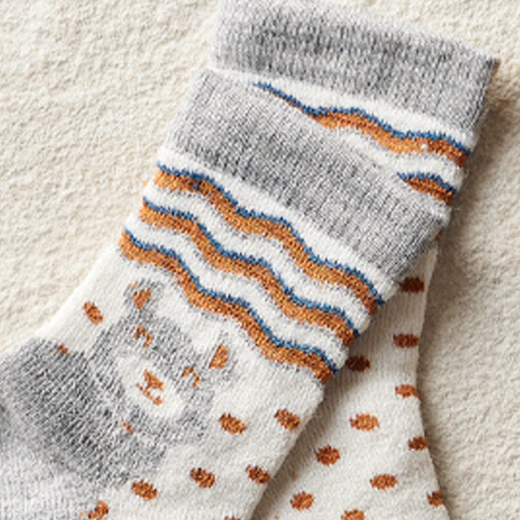 A pair of gray and white knit socks with orange and blue wavy stripes and a gray cat face motif, laid flat on a textured beige surface.