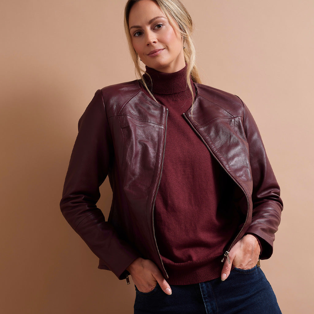 A woman wearing a dark burgundy leather jacket over a matching burgundy turtleneck sweater and dark blue jeans, posing against a solid beige background.