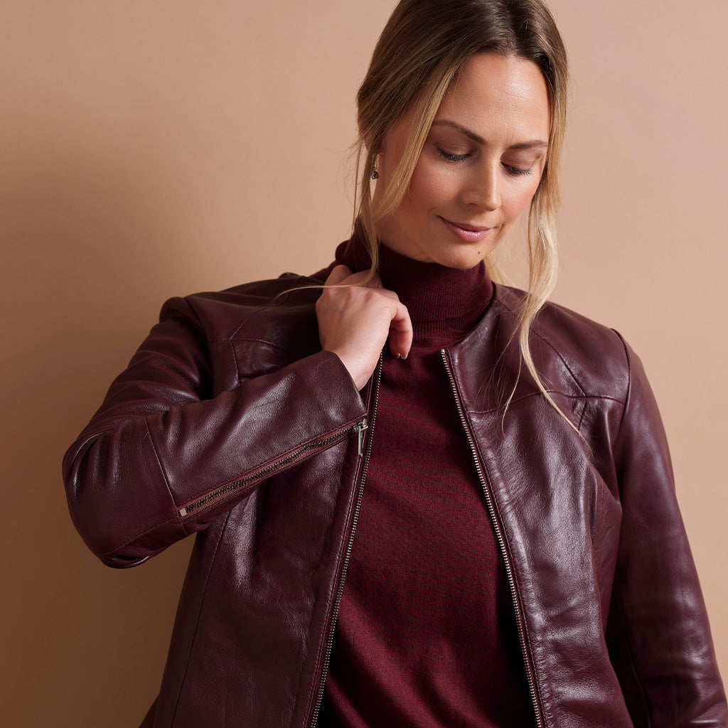 A woman wearing a dark burgundy leather moto jacket over a matching burgundy turtleneck sweater, posed against a solid warm beige background.