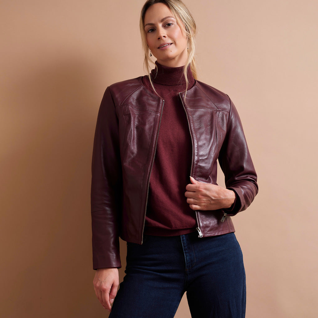 A woman modeling a dark burgundy leather moto jacket over a matching burgundy turtleneck sweater and dark blue jeans, set against a solid warm beige studio background.