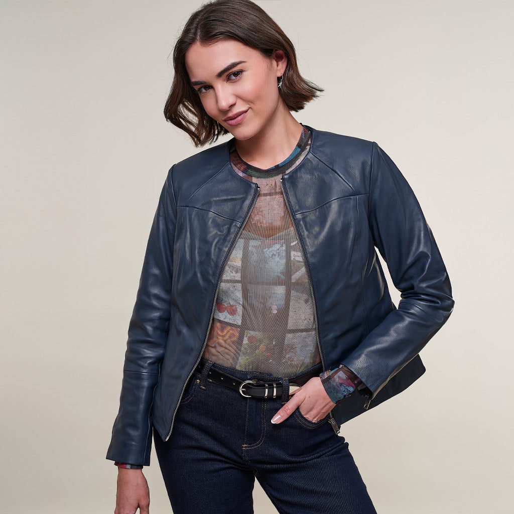A woman modeling a dark navy blue leather jacket over a patterned top and dark jeans, photographed against a plain light beige studio background.